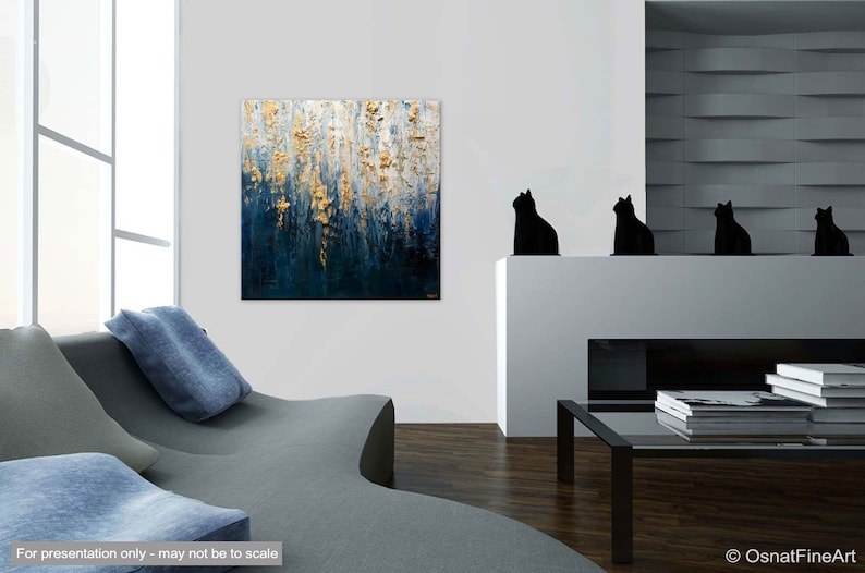 May include: Abstract painting with gold leaf accents over a dark blue and white background. The artwork is displayed in a modern living room setting, with a gray sofa and black cat figurines on a white mantel. The painting is signed in the bottom right corner.