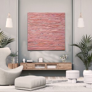 May include: A modern living room interior with a pink abstract painting hanging on the wall above a wooden media console. The room features a white armchair, a white ottoman, two white plant stands with potted plants, and a white rug.