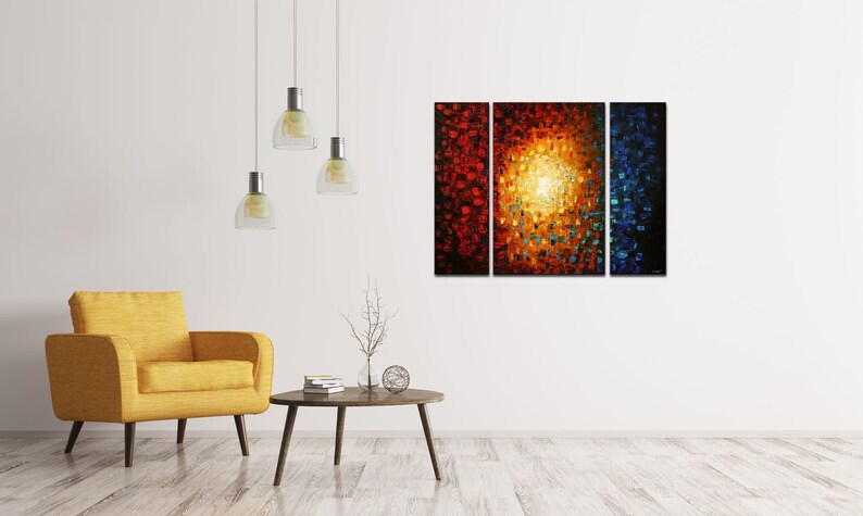 May include: A three-piece abstract painting with a bright yellow center surrounded by red, orange, and blue hues. The painting is displayed on a white wall in a living room setting with a yellow armchair and a coffee table.