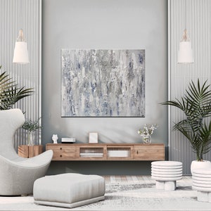 May include: A modern living room interior with a gray and white abstract painting hanging on the wall above a wooden media console. A gray armchair with a matching ottoman sits in front of the console. Two potted palm trees flank the console, and two white stacked side tables are in the foreground.