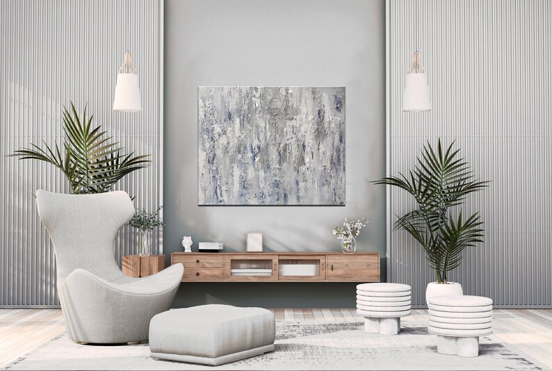 May include: A modern living room interior with a gray and white abstract painting hanging on the wall above a wooden media console. A gray armchair with a matching ottoman sits in front of the console. Two potted palm trees flank the console, and two white stacked side tables are in the foreground.