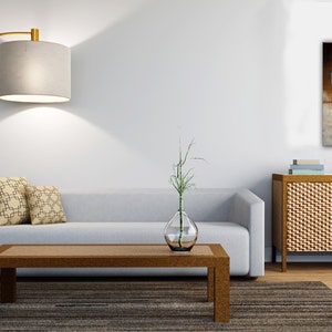 May include: A light gray sofa with two patterned pillows sits in a living room with a wooden coffee table, a gold floor lamp, a vase with a green plant, and a wooden cabinet with a brown abstract painting above it.