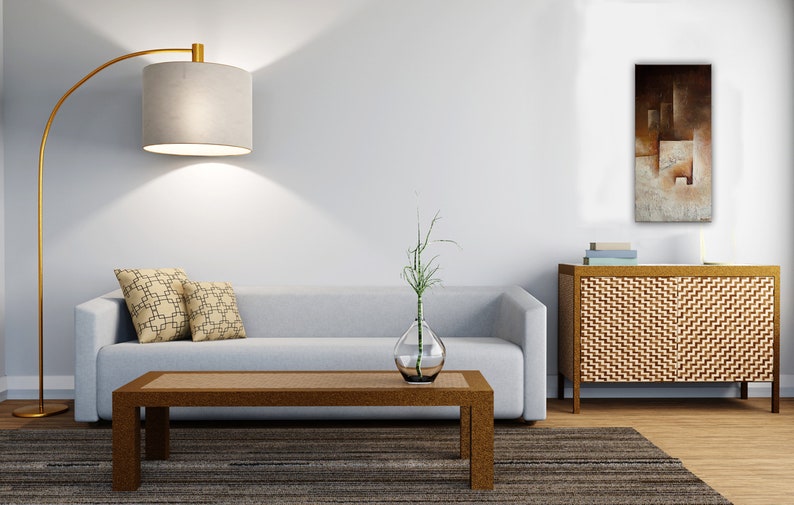 May include: A light gray sofa with two patterned pillows sits in a living room with a wooden coffee table, a gold floor lamp, a vase with a green plant, and a wooden cabinet with a brown abstract painting above it.