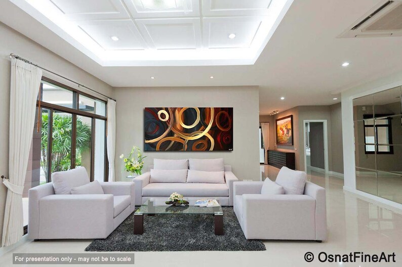 May include: Abstract painting with gold, red, and black circles on a black background. The painting is hanging on a wall in a living room with white couches and a coffee table.