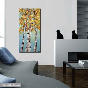 May include: Abstract painting of three birch trees with yellow and gold leaves against a blue background. The painting is framed and hanging on a white wall in a living room setting.