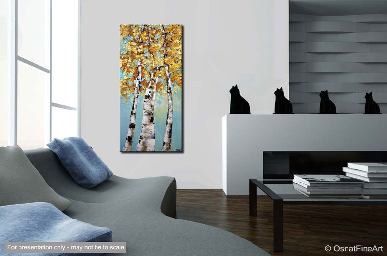 May include: Abstract painting of three birch trees with yellow and gold leaves against a blue background. The painting is framed and hanging on a white wall in a living room setting.