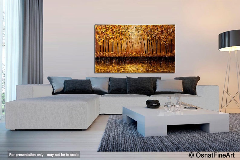 May include: A large abstract painting of a forest scene with golden and brown hues. The painting depicts a group of trees with their branches reaching towards the sky. The light shines through the trees, creating a warm and inviting atmosphere. The painting is displayed on a white wall above a white couch and coffee table.
