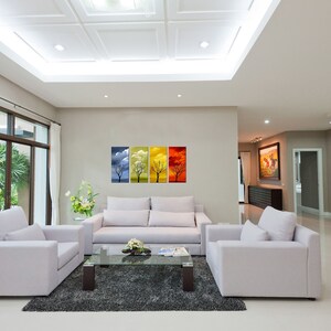 May include: A living room interior with white walls and a white ceiling. There are two white armchairs and a white sofa in the room. A glass coffee table sits in front of the sofa. A set of four framed prints depicting trees in different seasons hangs on the wall. The prints are blue, yellow, orange, and red.