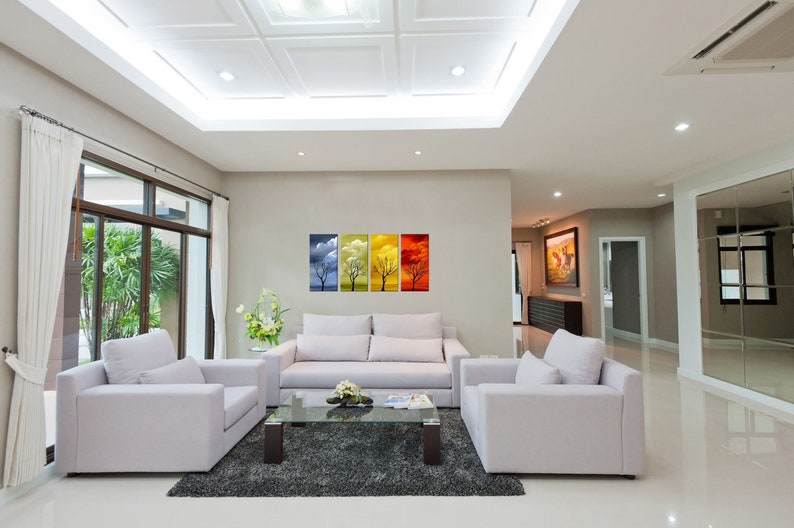 May include: A living room interior with white walls and a white ceiling. There are two white armchairs and a white sofa in the room. A glass coffee table sits in front of the sofa. A set of four framed prints depicting trees in different seasons hangs on the wall. The prints are blue, yellow, orange, and red.