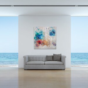 May include: Abstract painting with blue, red, yellow, and white colors on a white canvas. The painting is hanging on a white wall in a modern living room with a gray sofa and large windows overlooking the ocean.