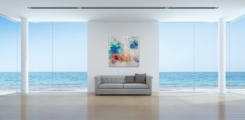 May include: Abstract painting with blue, red, yellow, and white colors on a white canvas. The painting is hanging on a white wall in a modern living room with a gray sofa and large windows overlooking the ocean.