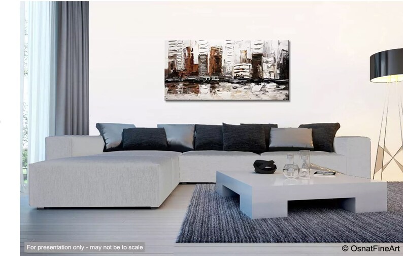 May include: A white, gray, and brown abstract painting of a cityscape with a reflection of the buildings in the water below. The painting is hanging on a white wall above a gray sectional sofa with black pillows. A white coffee table sits in front of the sofa.