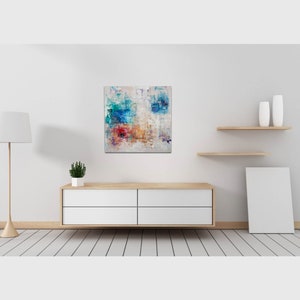 May include: Abstract painting with blue, red, yellow, and white paint on a white canvas. The painting is hanging on a white wall above a wooden dresser with four drawers.