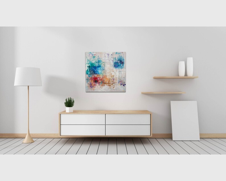 May include: Abstract painting with blue, red, yellow, and white paint on a white canvas. The painting is hanging on a white wall above a wooden dresser with four drawers.