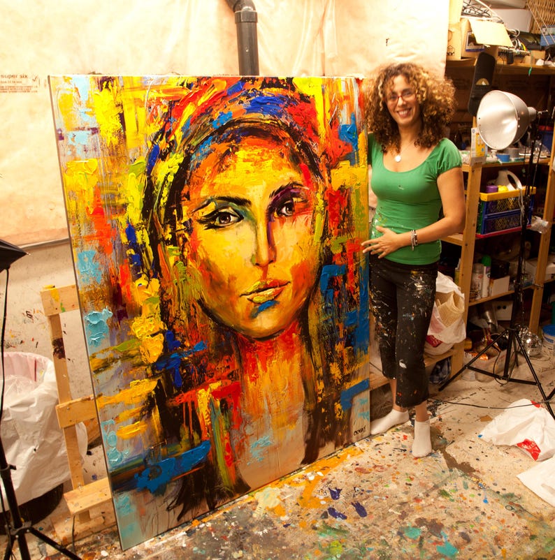 May include: A large, colorful abstract portrait painting of a woman with a yellow and orange face, blue and yellow head covering, and red and blue accents. The painting is on an easel.