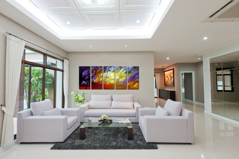 May include: A living room interior with a white sofa and two armchairs. A large abstract painting of a sunset with clouds and a tree hangs on the wall. The painting is in five panels.