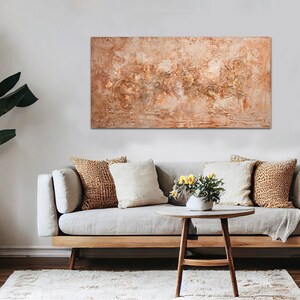 May include: An abstract textured painting in warm tones of beige, brown, and gold hangs above a light gray sofa. The living room setting includes decorative pillows, a wooden coffee table, and a large potted plant in a woven basket.