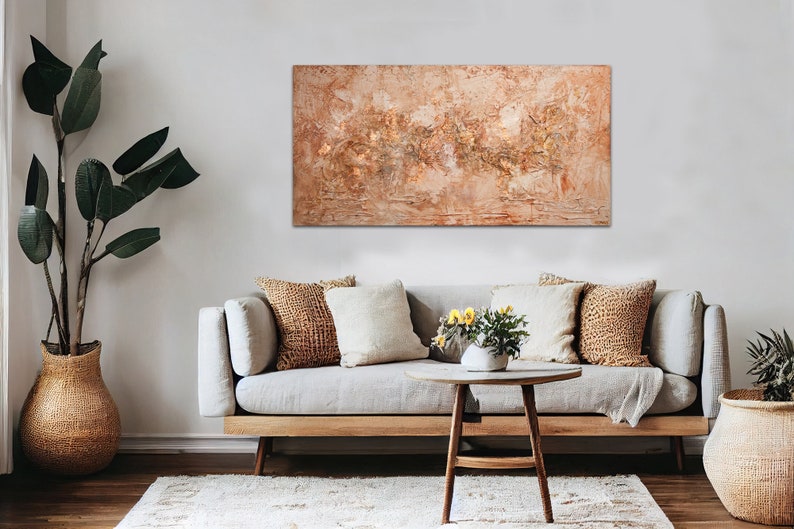 May include: An abstract textured painting in warm tones of beige, brown, and gold hangs above a light gray sofa. The living room setting includes decorative pillows, a wooden coffee table, and a large potted plant in a woven basket.