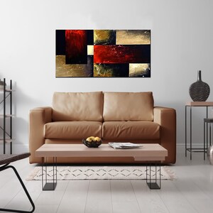 May include: Abstract painting with geometric shapes in shades of red, gold, and black. The painting is displayed on a wall in a living room setting with a brown leather sofa, a coffee table, and decorative items.