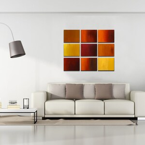 May include: A white couch with three pillows in a living room setting. A floor lamp with a gray shade is in the corner. A nine-piece abstract art piece with red, orange, and yellow squares is hanging on the wall. A potted plant is in the corner of the room.