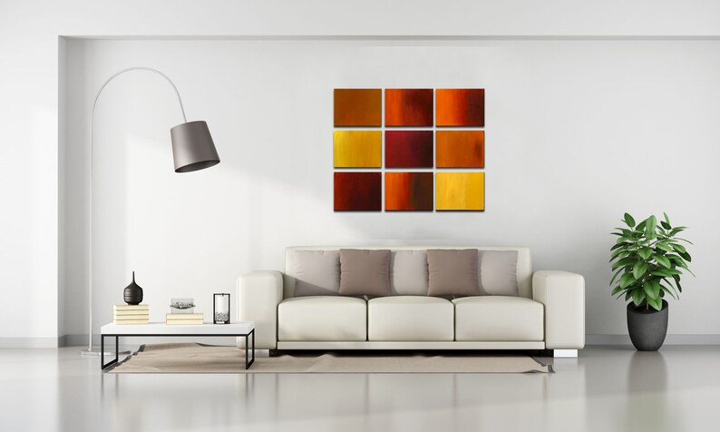 May include: A white couch with three pillows in a living room setting. A floor lamp with a gray shade is in the corner. A nine-piece abstract art piece with red, orange, and yellow squares is hanging on the wall. A potted plant is in the corner of the room.