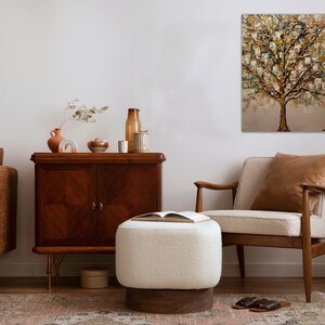 May include: A living room interior design with a brown leather sofa, a white ottoman, a wooden armchair, a wooden cabinet, and a painting of a gold tree on the wall.