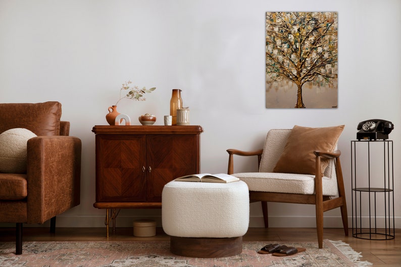 May include: A living room interior design with a brown leather sofa, a white ottoman, a wooden armchair, a wooden cabinet, and a painting of a gold tree on the wall.
