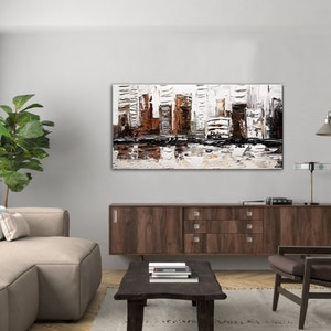 May include: A modern living room interior with a brown wooden cabinet, a beige sofa with pillows, a brown armchair, a coffee table with books, a potted plant, and a large abstract painting of a city skyline in shades of white, brown, and black.