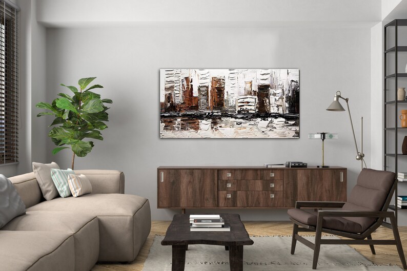 May include: A modern living room interior with a brown wooden cabinet, a beige sofa with pillows, a brown armchair, a coffee table with books, a potted plant, and a large abstract painting of a city skyline in shades of white, brown, and black.