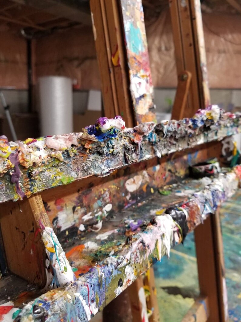 May include: A wooden artist's easel covered in dried paint. The paint is a variety of colours, including white, blue, green, yellow, and red. The easel has a shelf for holding paint tubes or brushes.