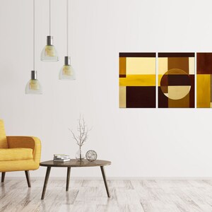 May include: A three-piece abstract art print set featuring geometric shapes in shades of brown and yellow. The artwork is displayed on a white wall in a living room setting with a yellow armchair and a coffee table.