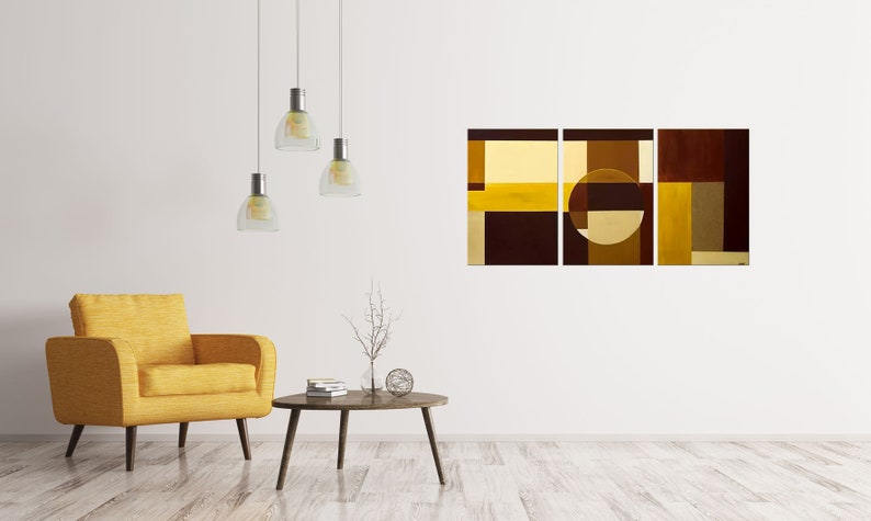 May include: A three-piece abstract art print set featuring geometric shapes in shades of brown and yellow. The artwork is displayed on a white wall in a living room setting with a yellow armchair and a coffee table.