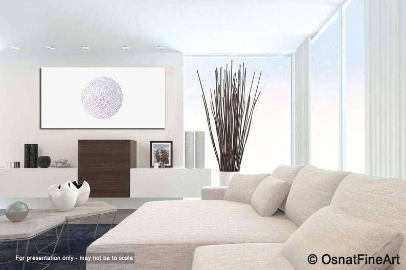 May include: White textured circle abstract painting on a white canvas, hanging on a white wall in a modern living room with a white couch and a vase of brown branches.