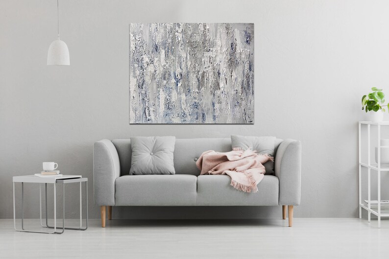 May include: A gray sofa with a pink throw blanket and a gray pillow in a living room with a white wall and a painting above the sofa.