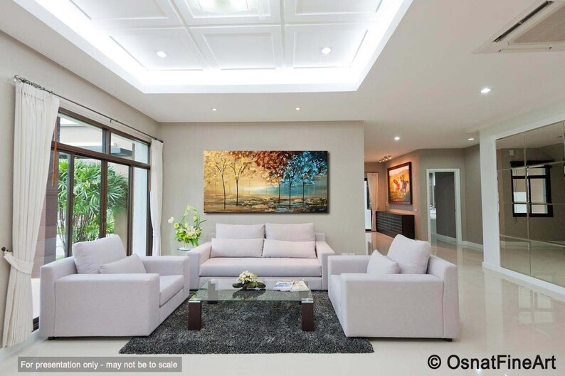 May include: A living room interior design featuring a large abstract painting of trees in shades of gold, red, blue, and green. The painting is hung on a wall above a white sofa and two white armchairs. The room has a white ceiling with recessed lighting and a large window with white curtains.