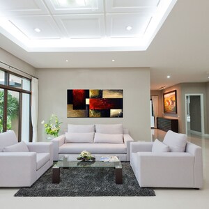 May include: A modern living room with white walls and a white sofa. There is a large abstract painting on the wall behind the sofa. The painting is in shades of red, brown, and gold. There are two white armchairs in the room, and a glass coffee table in front of the sofa.