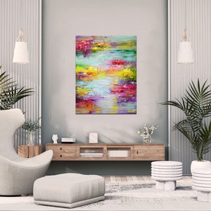 May include: Abstract painting with vibrant colors of pink, yellow, green, and blue. The painting is displayed on a wall in a modern living room with a white chair, a wooden shelf, and two white plant stands with potted plants.