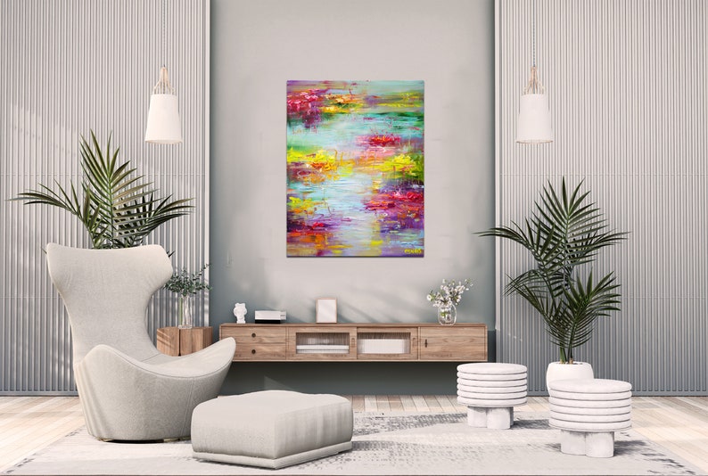 May include: Abstract painting with vibrant colors of pink, yellow, green, and blue. The painting is displayed on a wall in a modern living room with a white chair, a wooden shelf, and two white plant stands with potted plants.
