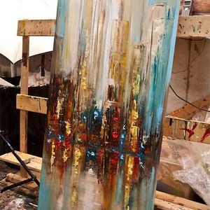 May include: Abstract painting on canvas with a cityscape theme. The painting features a mix of blues, browns, yellows, and reds. The painting is leaning against a wooden easel.
