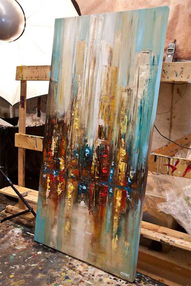 May include: Abstract painting on canvas with a cityscape theme. The painting features a mix of blues, browns, yellows, and reds. The painting is leaning against a wooden easel.