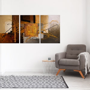 May include: A three-piece abstract art print set featuring a gold, brown, and black color scheme. The artwork is displayed on a white wall above a gray armchair and a white side table.