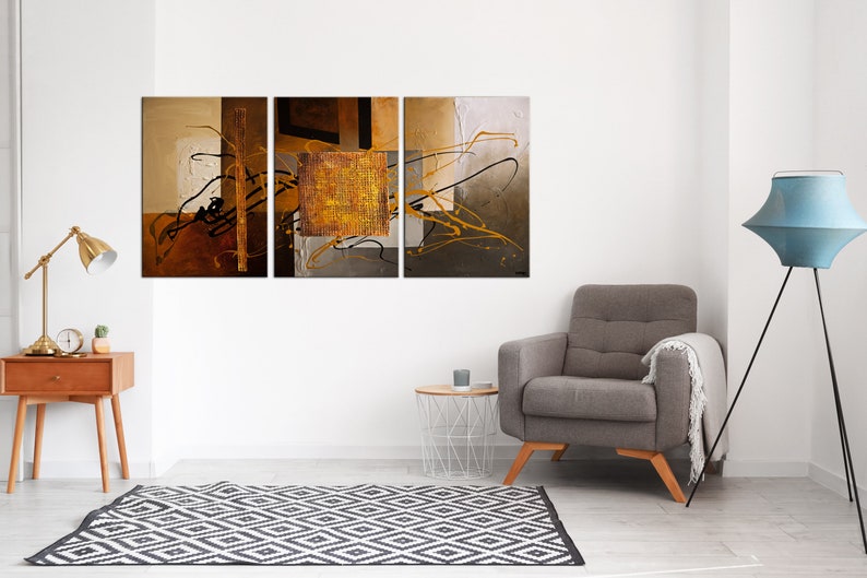 May include: A three-piece abstract art print set featuring a gold, brown, and black color scheme. The artwork is displayed on a white wall above a gray armchair and a white side table.
