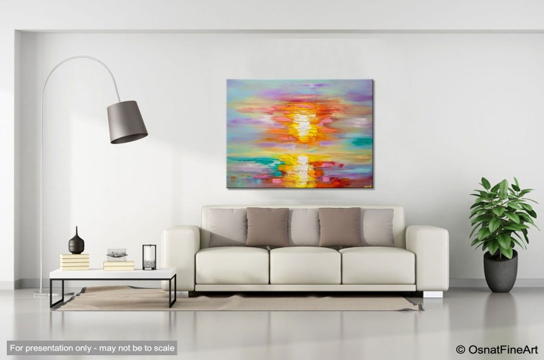 May include: Abstract painting of a sunset over water with vibrant colors of pink, orange, yellow, blue, and green. The painting is displayed on a white wall above a white sofa with gray pillows.