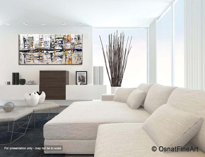 May include: Abstract painting with a white background and colorful geometric shapes. The painting is hanging on a white wall in a modern living room with a white couch and a coffee table.