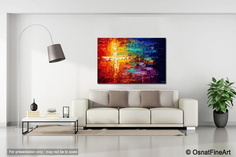 May include: Abstract painting with a white cross in the center, surrounded by vibrant colors like red, orange, yellow, green, blue, and purple. The painting is displayed on a white wall above a white sofa with pillows.