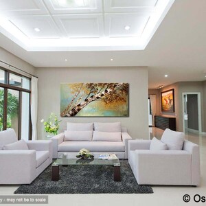 May include: A living room interior with a large abstract painting of a tree with brown and gold leaves hanging on a light beige wall. The painting is above a white sofa with two matching white armchairs. The room has a white ceiling with recessed lighting and a large window with white curtains.