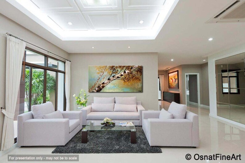 May include: A living room interior with a large abstract painting of a tree with brown and gold leaves hanging on a light beige wall. The painting is above a white sofa with two matching white armchairs. The room has a white ceiling with recessed lighting and a large window with white curtains.