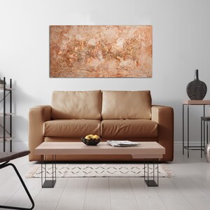 May include: An abstract painting with a textured surface in shades of beige, brown, and gold hangs above a tan leather sofa. The living room features a wooden coffee table, black metal side tables, and decorative vases.