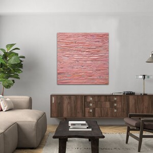 May include: A pink abstract painting with horizontal lines and a textured surface hangs on a white wall above a brown wooden cabinet. The painting is in a modern living room setting with a sofa, armchair, and coffee table.