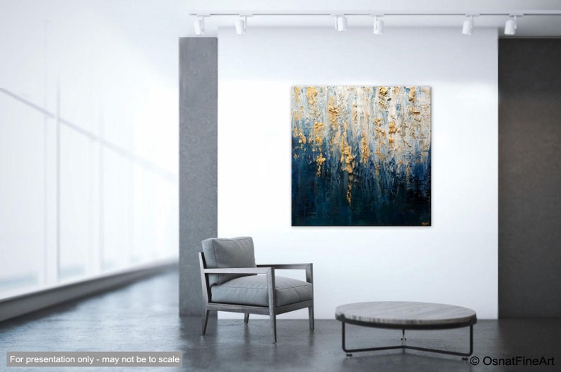 May include: Abstract textured painting with gold and deep blue tones. The artwork is displayed on a white wall in a modern interior setting. A gray armchair and a round coffee table are in the foreground.